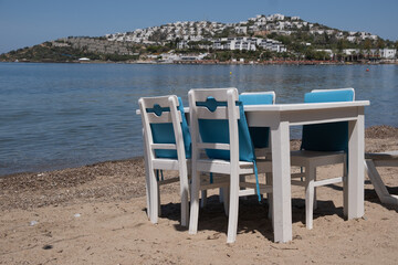 Fototapeta premium Table and chairs on sandy beach near sea with background of white buildings on hill