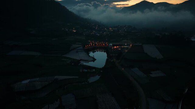 Aerial view of Dieng Plateau with village lights reflecting in a lake surrounded by agricultural fields and misty mountains at night in Kejajar, Central Java, Indonesia.