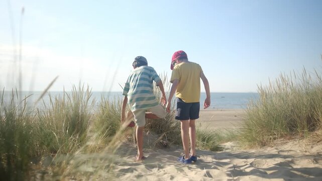 Back view of two young boys standing on a sandy path between dunes, emptying their shoes of sand before running towards the calm ocean for a summer adventure