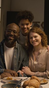 Vertical front view slow motion shot of multiethnic family posing together at dinner table all looking at camera with smiles, teen son embracing parents