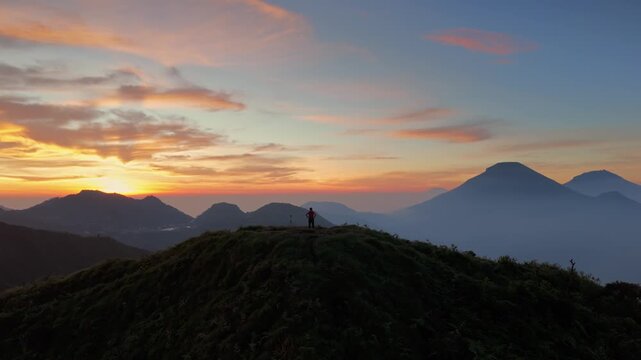 Aerial view of a person standing on a grassy mountain ridge overlooking volcanic peaks and mist during sunrise in Kejajar, Central Java, Indonesia.