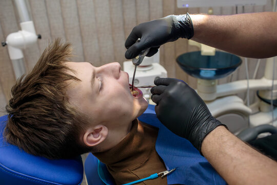  teenage boy lying in blue dental chair with eyes closed while dentist in black gloves uses metal instrument inside open mouth at clinic