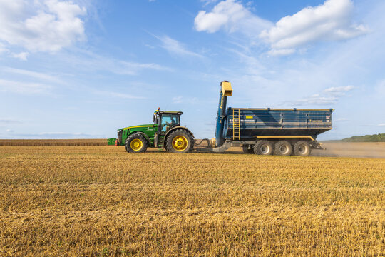 Lutsk, Ukraine &ndash; August 7, 2025: A tractor pulls a trailer while harvesting crops in a large field. Dust rises as they work under a clear sky, showing the farming activity