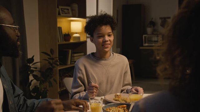 Medium shot of smiling Black teenage boy enjoying meal with multiracial family sitting at dinner table together at home, scene lit with warm evening lighting