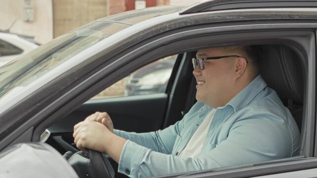 Man seated in car with hands on steering wheel smiling and gently bobbing while gripping the wheel, visible dashboard and parked cars on street; contentment.