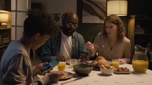 Side view shot of Black teenage boy using smartphone at dinner table and showing funny video to smiling parents sharing interests, copy space