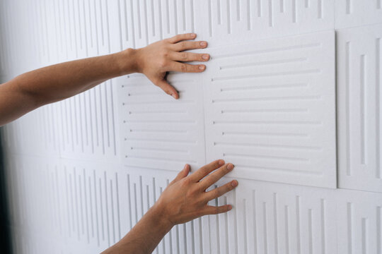 Hands installing soundproofing acoustic panels on a white studio wall, creating a quiet environment for sound dampening and noise reduction in a recording space.