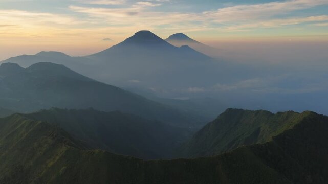 Aerial view of the Dieng Plateau featuring misty volcanic peaks and green mountain ridges under a soft golden sunrise sky in Kejajar, Central Java, Indonesia.