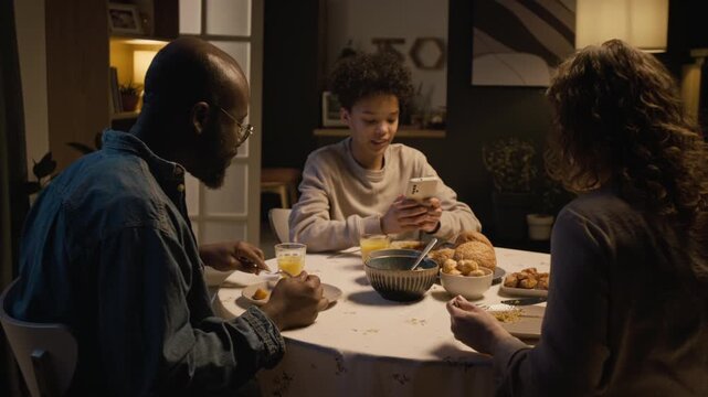 Multiracial family sitting at dinner table together at home and sharing meal with focus on Black teenage boy using smartphone, copy space