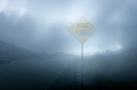 A yellow diamond shaped traffic sign stating Fog Area in very heavy fog with traffic in the distance