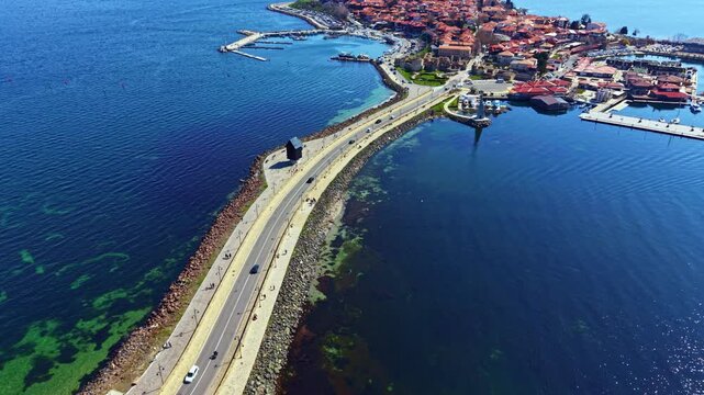 People walk along a road next to the water near Nesebar Old Town. Boats are docked in the harbor. The skyline of the town shows buildings and rooftops under clear blue skies.