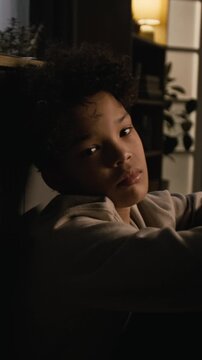 Vertical side view shot of upset African American boy sitting on floor in dark room and turning looking at camera, mental health issues in adolescents, story format