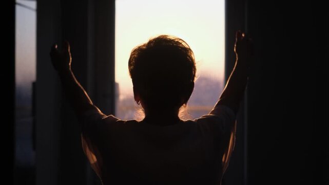 An old woman walks towards the light with her hands raised, seen from behind