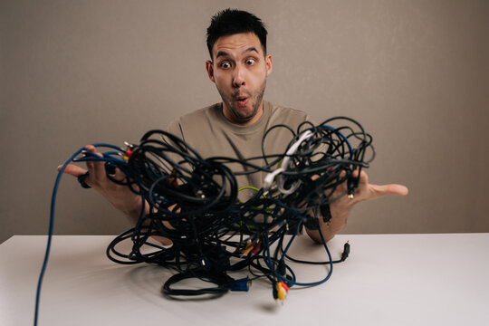 Studio portrait of frustrated man expressing anger and irritation while struggling with a chaotic pile of tangled cables, symbolizing technology problems and digital stress.