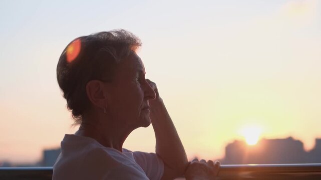 An elderly woman contemplates the sunset while standing on a balcony