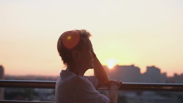 A pensive old woman standing on a balcony at sunset