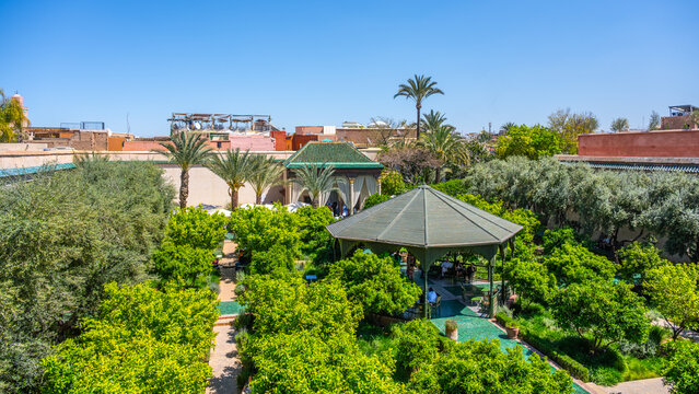 Visitors walk through the Secret Garden in Marrakesh, Morocco, surrounded by greenery. Palm trees and orange trees fill the landscape under a clear blue sky.
