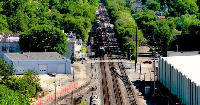 Aerial view of freight trains on multiple tracks passing through industrial areas and lush green trees in Cincinnati, Ohio, United States.