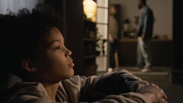 Side view close up shot of upset Black boy sitting on floor in dark room at home listening to parents fighting in blurred background, copy space