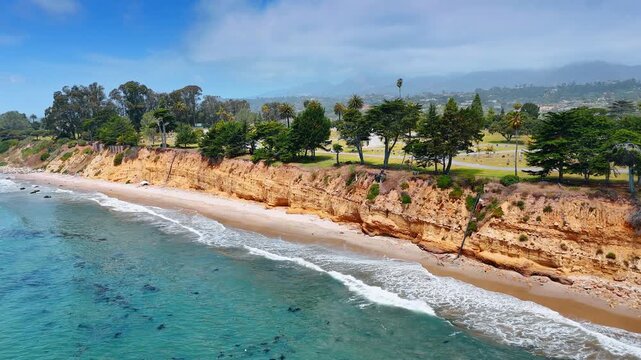Waves roll to the sandy beach along the cliffs with green trees growing on. Coast of the Pacific Ocean in Santa Barbara, California, USA.