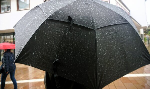 R&uuml;ckenansicht von stehendem Mann mit schwarzem aufgespannten Regenschirm mit Wassertropfenmuster auf Stra&szlig;e in Stadt bei Regen am Nachmittag im Herbst