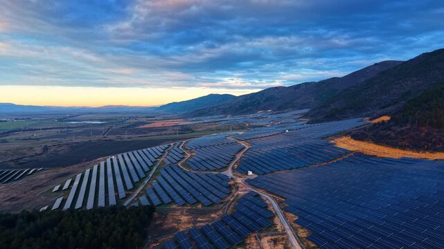 Flying above the field of photovoltaic cells near the mountains. Dramatic cloudscape covers the sky over the scenery at sunset.