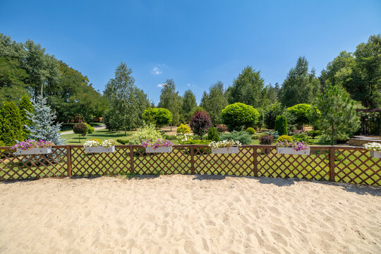 Wooden lattice fence decorated with rectangular flower planter boxes separates a sandy area from a landscaped garden area containing various green trees and shrubs under a clear blue sky