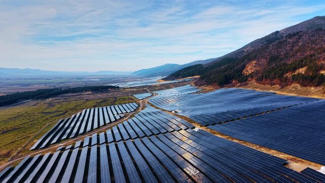 Glowing modern solar panels in the long rows. Drone footage over the field of photovoltaic cells near the mountains.