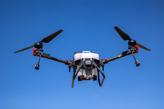 An unmanned aerial vehicle configured for agricultural spraying operations hovers in a clear blue sky with its dual black propellers in motion above its white liquid payload storage tank