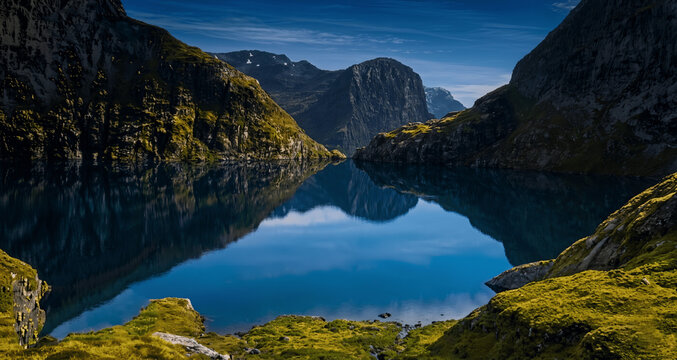 Tranquil Fjords Reflection: A serene alpine lake mirrors the dramatic, moss-covered mountains under a clear blue sky, evoking a sense of peace and solitude in a pristine natural landscape.