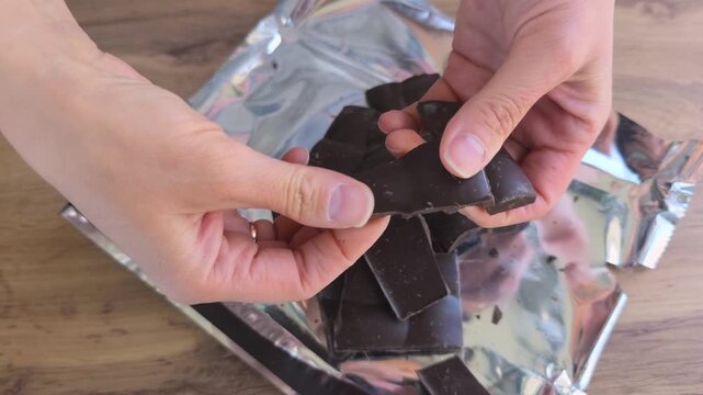 Hands of a person breaking dark chocolate pieces from a foil wrapper, showcasing the continuous action of separating chocolate squares on a wooden surface