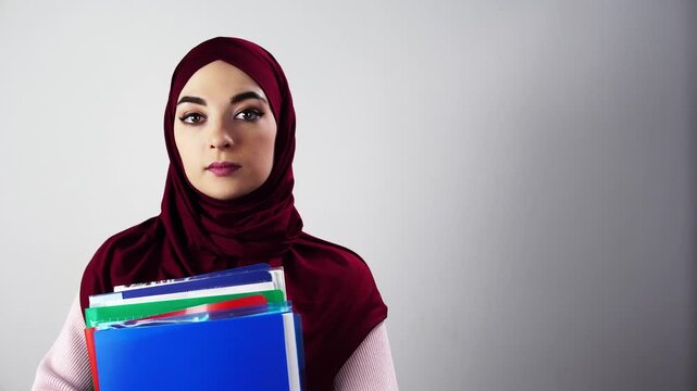 Portrait of woman in hijab holding folders and documents against neutral background, confident professional stance, clear direct gaze and neat presentation for corporate, academic, or recruitment