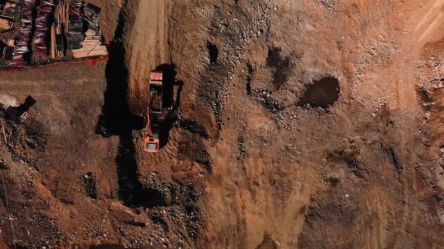 Excavator is engaged in trenching on steep incline revealing soil stratification. Aerial view of trenching scene with truck. Slope and bucket scars are localized in erosion pockets and muddy puddles 