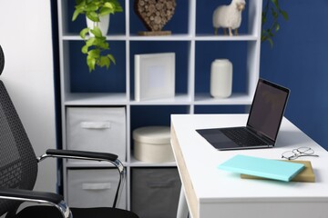 Fototapeta premium White desk with laptop, stationery, chair, shelving unit and decor in office. Interior design