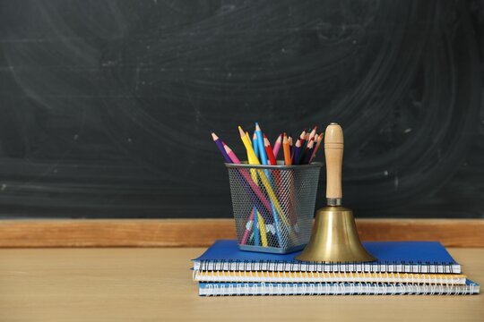 School holidays. Golden bell, notebooks and pencils on wooden table near chalkboard, closeup. Space for text