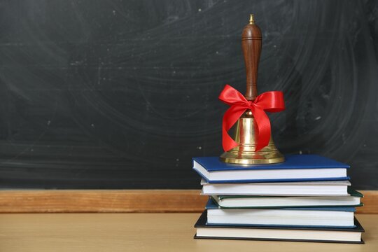 School holidays. Golden bell and books on wooden table near chalkboard, closeup. Space for text
