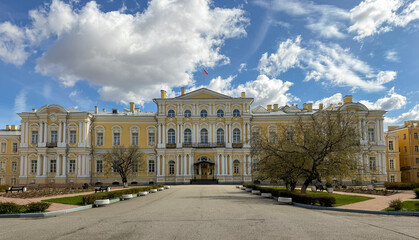 Naklejka premium Historic yellow building features classical architecture and large windows, surrounded by trees and a paved pathway under a blue sky with clouds