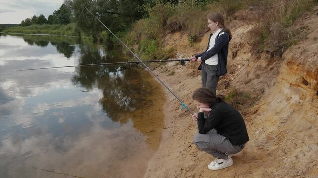 Two people fishing riverbank sandy shore, quiet rural setting with mentor guiding teenager, casting rods and checking reel, reflective water mirrors sky and trees, warm summer light, casual jackets