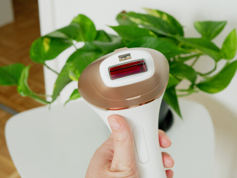 Female Hand Holding IPL Laser Hair Removal Device Front View of Red Flash Lamp, Home Beauty Skin Care Technology with Green Plant Background