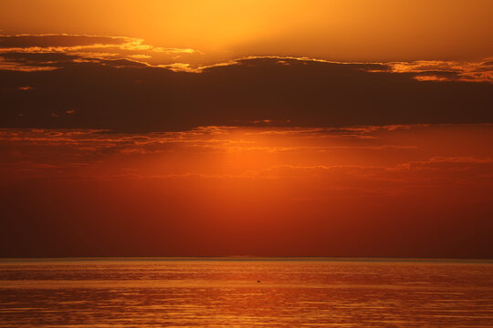 Dramatic evening sky over the sea. The setting sun hides behind heavy dark clouds, casting a fiery orange reflection on the calm water surface.