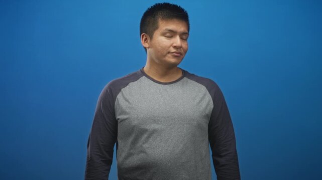 Young man in gray raglan shirt turns head right with hands mid gesture in blue studio; thoughtful introspection.