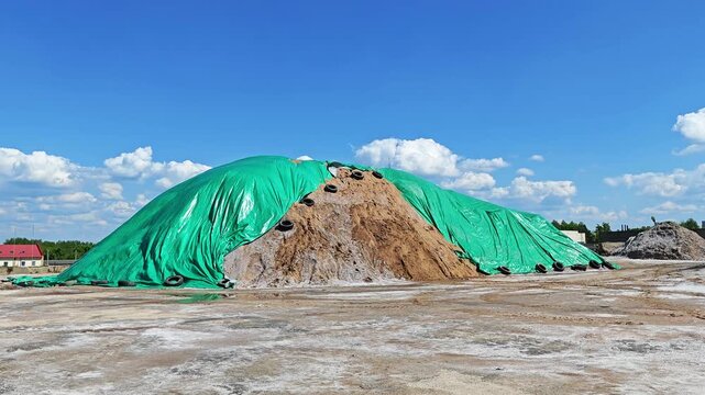 Huge sand-salt pile for winter road maintenance covered with green PVC film against rain, blowing in the wind under blue sky. 4K footage of seasonal deicing material storage.