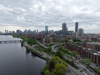 Obraz premium Aerial drone view of the Boston skyline under a cloudy sky, featuring the Charles River, Harvard Bridge, and Back Bay brownstones leading toward the Prudential and John Hancock towers.