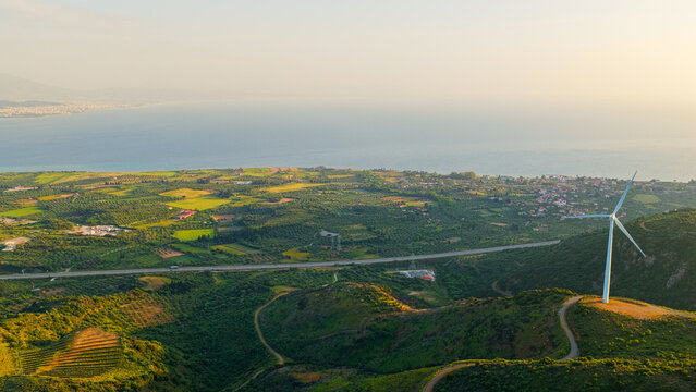 Wind turbine standing on a hill, generating renewable electricity against a backdrop of green landscape, a coastal road, and the vast blue sea under a clear sky