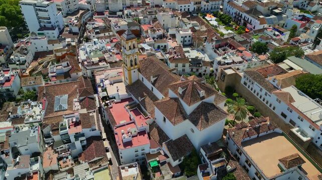 Aerial orbit view of the historic Encarnacion Church in the Old Town of Marbella. Beautiful Baroque architecture with a tall bell tower surrounded by traditional white buildings in Spain.