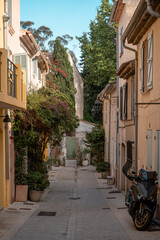 Obraz premium Narrow cobblestone street lined with pastel houses, potted plants, and parked scooter in Saint Tropez, France under clear blue sky.