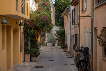 Obraz premium Narrow cobblestone street with yellow and peach buildings, potted plants, and a parked motorcycle in Saint Tropez, France, under natural daylight.