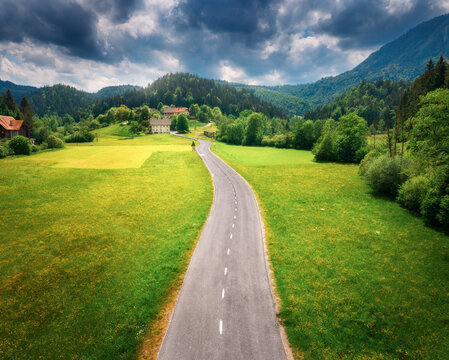 Aerial view of road in green meadows at sunset in summer. Top view from drone of rural road, mountains, forest. Beautiful landscape with roadway, sun rays, trees, hills, green grass, clouds. Slovenia