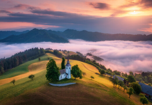 Aerial view of small church on the hill over pink low clouds at foggy sunrise in summer in Slovenia. Top view of beautiful chapel on mountain in fog, green meadows, trees, orange sky at dawn in spring