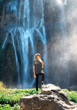 Young woman standing on a rock near big waterfall at sunset. Plitvice Lakes national park, Croatia. Lifestyle. Sporty girl on stone, waterfall. Female tourist enjoying nature, summer vacation. Travel